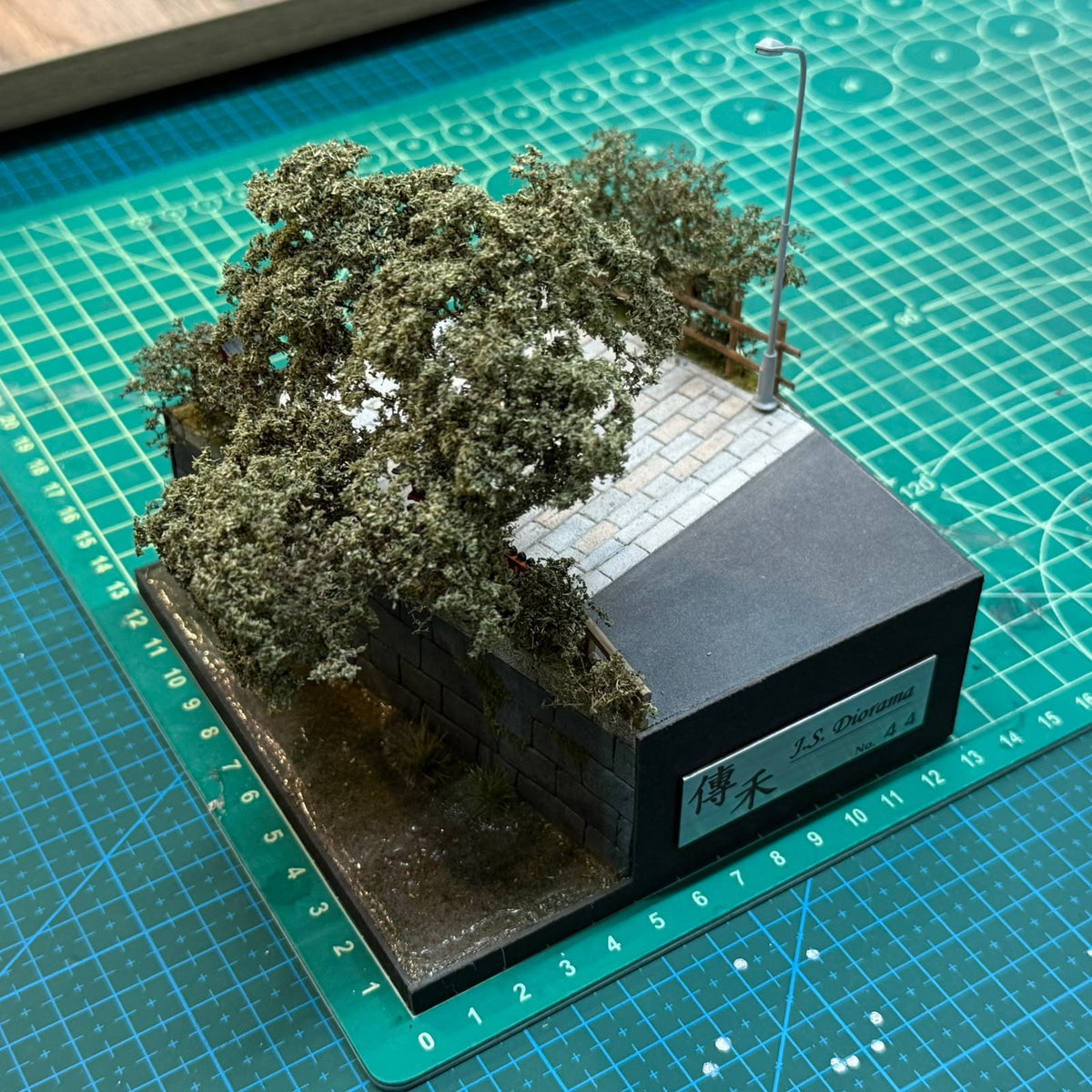 Miniature street diorama with stone base, dense trees, and a small lamp post