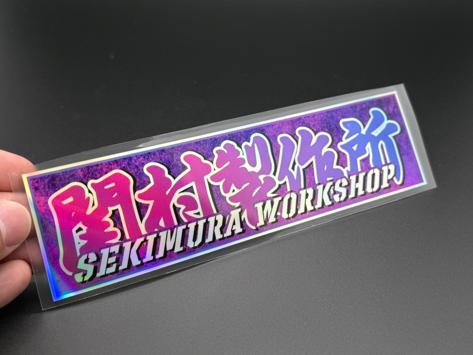 Hand-held view of purple rectangular sticker with Sekimura Workshop lettering, glossy