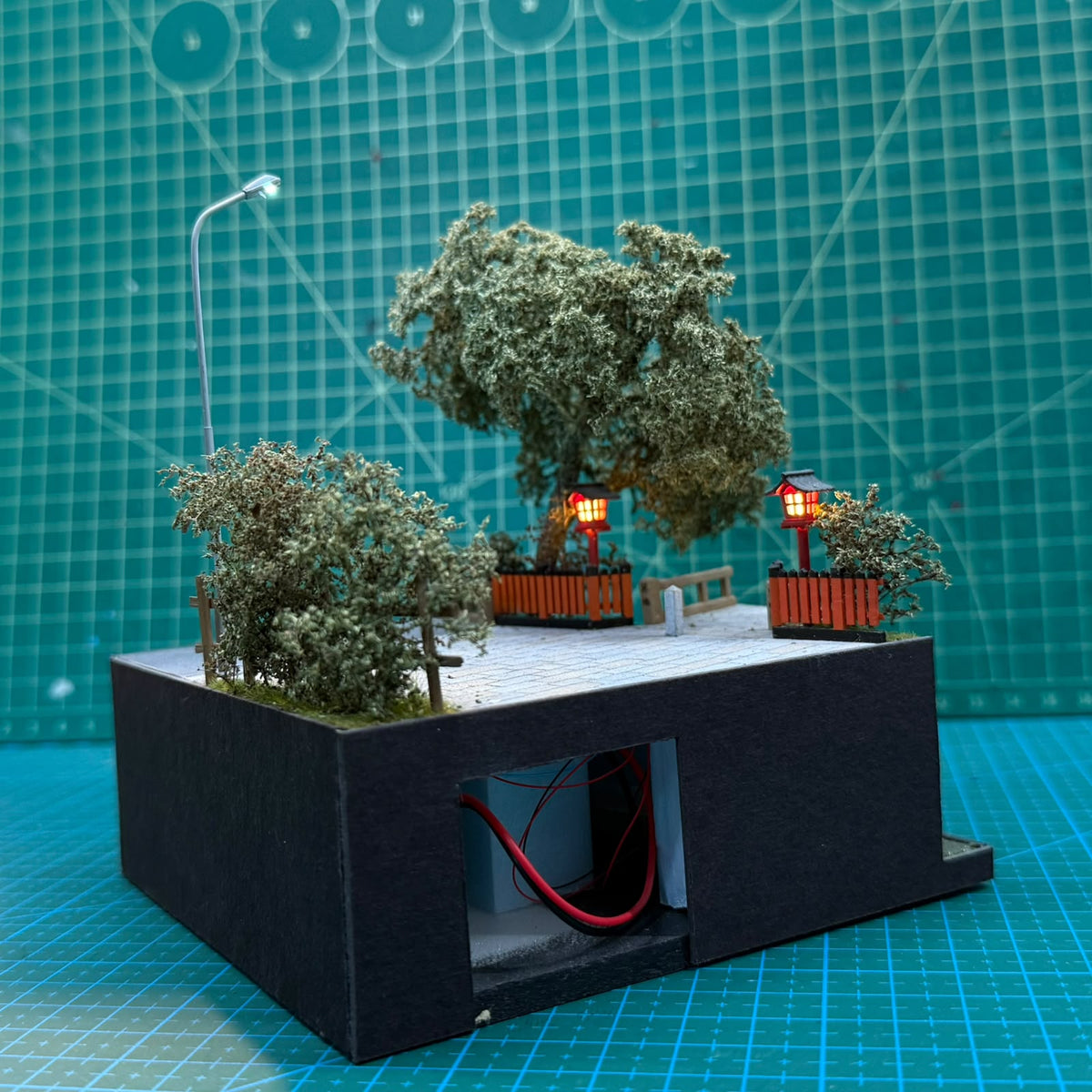 Miniature stone base diorama with lit red lantern posts, trees, and cable opening