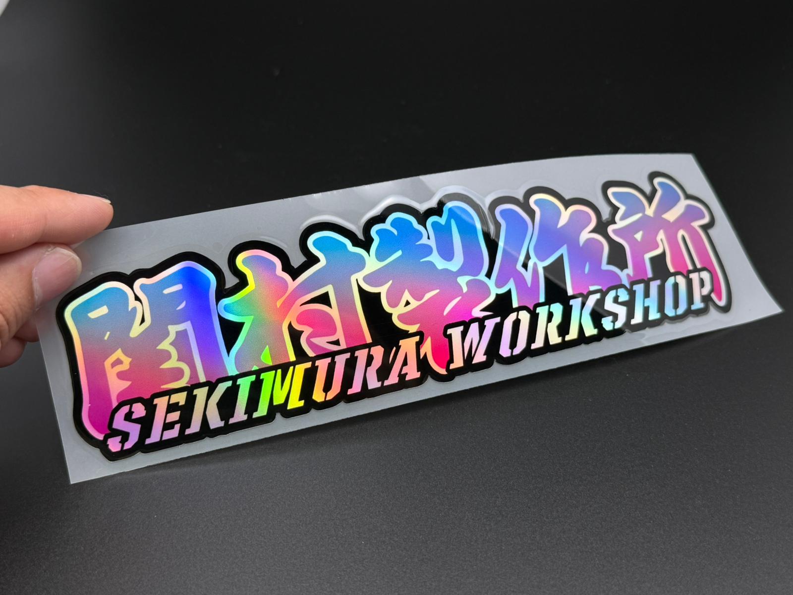 Sekimura Workshop sticker held in hand, blue and pink lettering on white backing