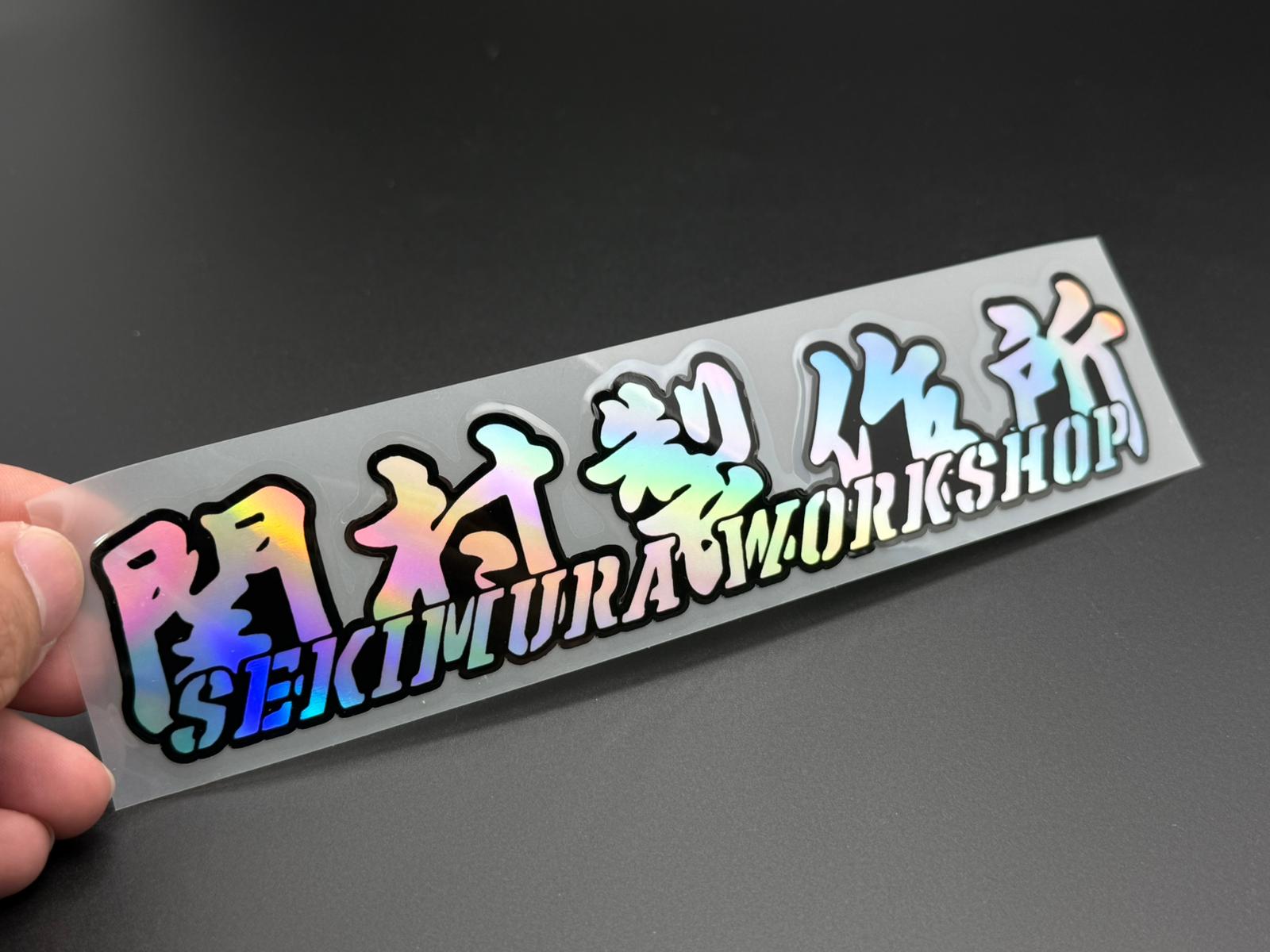 Hand-held graffiti-style sticker decal reading Sekimura Workshop, 20x4.2cm.
