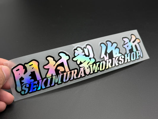 Hand-held graffiti-style sticker decal reading Sekimura Workshop, 20x4.2cm.